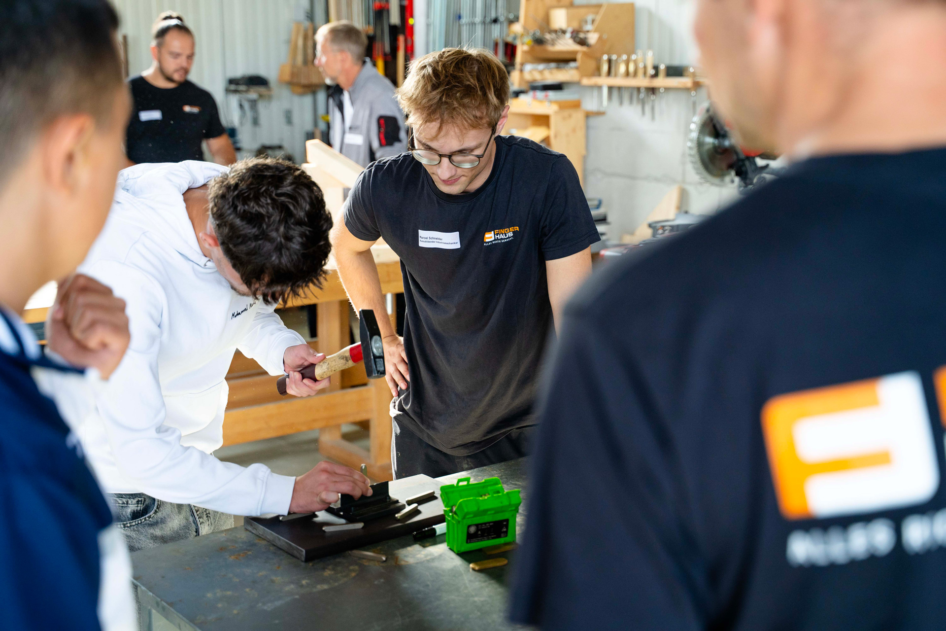 Mehrere junge Männer beobachten einen Mann mit Hammer und Meißel an einem Werkstück auf einem Werkstatttisch