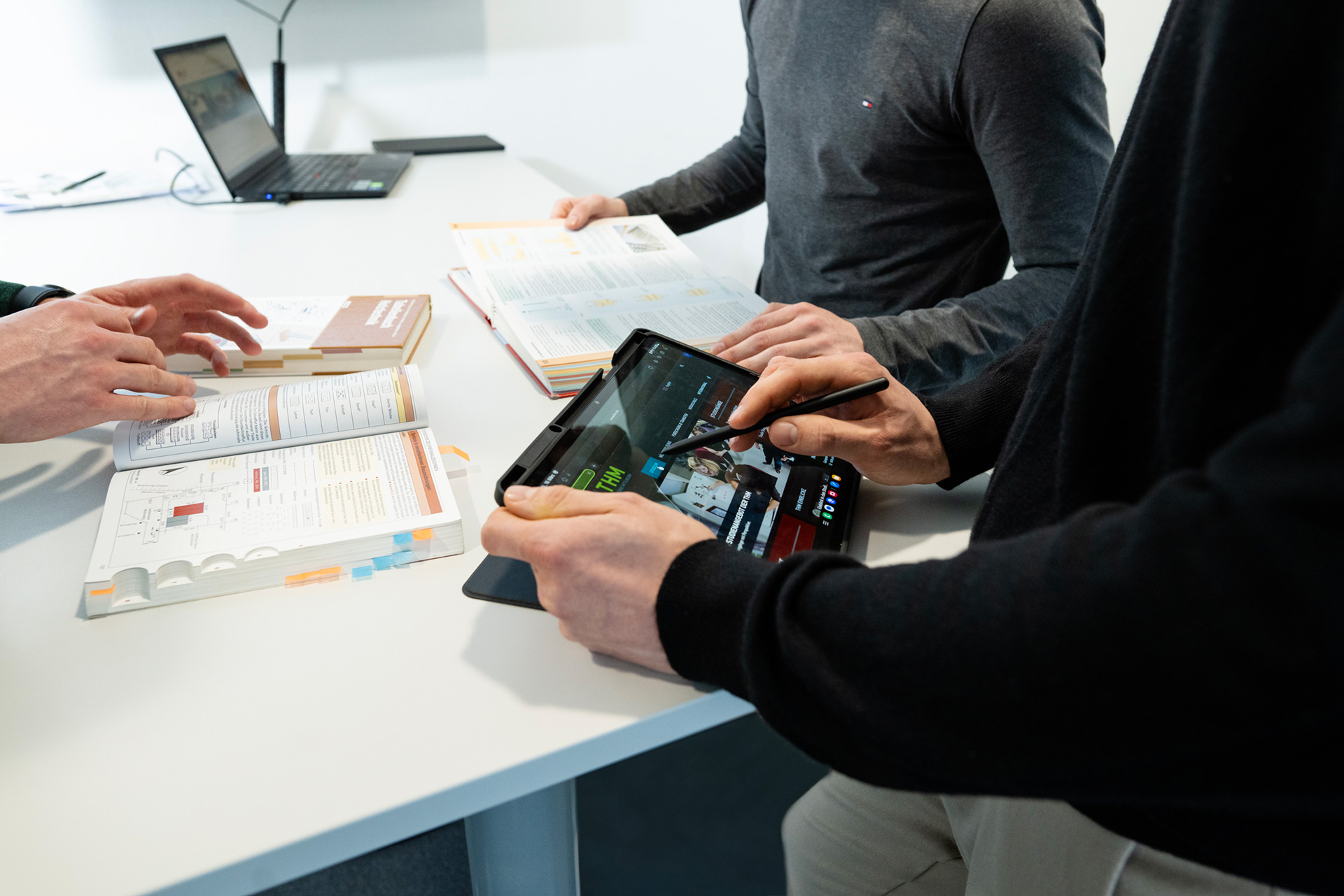 Zwei Personen stehen an einem weißen Tisch, eine hält einen Stift und bedient ein Tablet, die andere zeigt auf ein aufgeschlagenes Buch mit farbigen Markierungen
