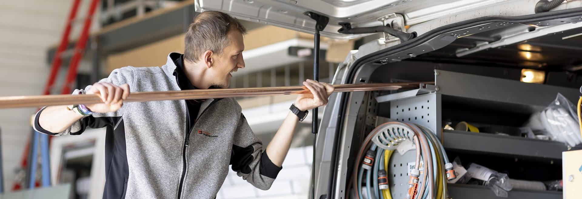Ein Mann lädt eine lange Metallstange in einen Transporter mit offenem Heck und Regalen voller Werkzeuge und Kabel ein