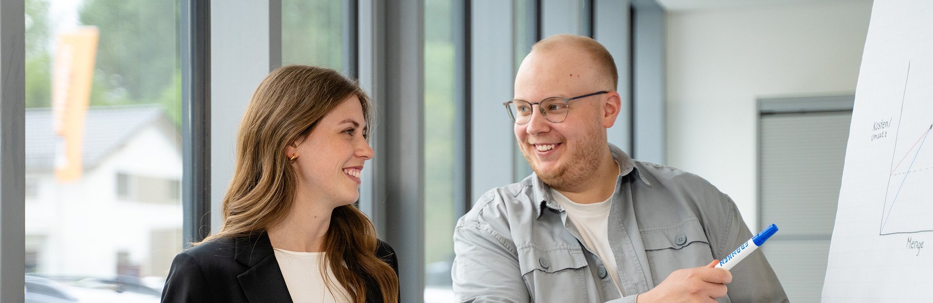 Zwei Personen stehen vor einem Flipchart, eine Person hält einen Marker und zeigt auf das Flipchart, die andere Person hört zu.