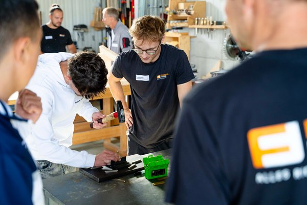 Mehrere junge Männer beobachten einen Mann mit Hammer und Meißel an einem Werkstück auf einem Werkstatttisch