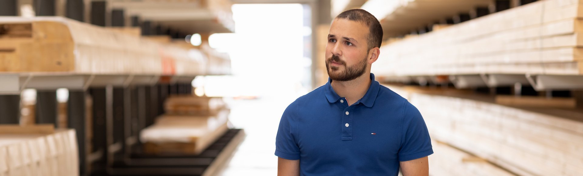 Mann mit kurzem Bart und blauem Poloshirt hält Klemmbrett und geht durch Lagerhalle mit Holzplattenregalen