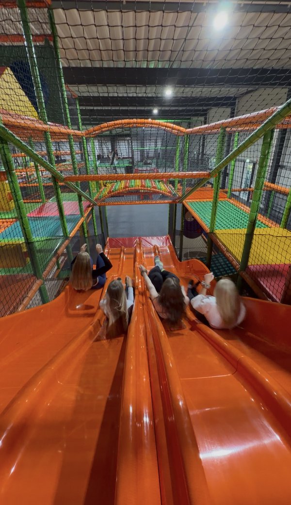 Vier Kinder rutschen nebeneinander auf orangenen Rutschen in einer bunten Indoor-Spielhalle