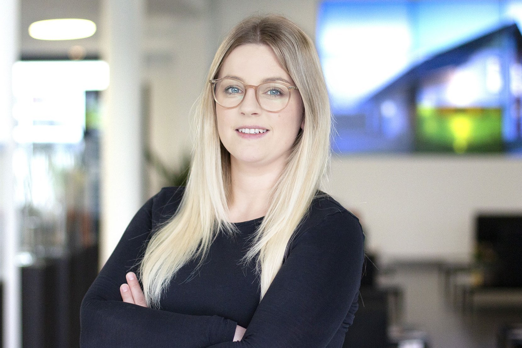 Frau mit langen blonden Haaren und schwarzem Pullover steht mit verschränkten Armen vor einem unscharfen modernen Bürohintergrund