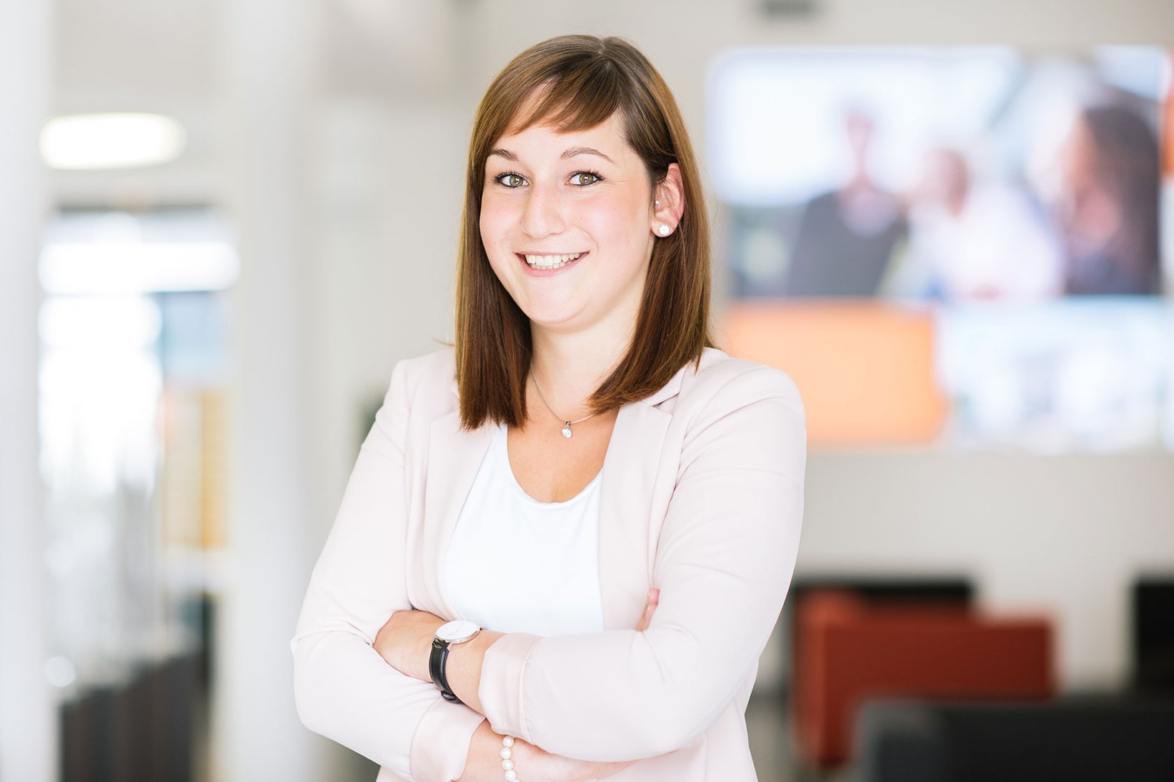 Frau mit schulterlangem Haar, weißem Oberteil und hellrosa Blazer steht mit verschränkten Armen in einem modernen Büro mit unscharfem Hintergrund.