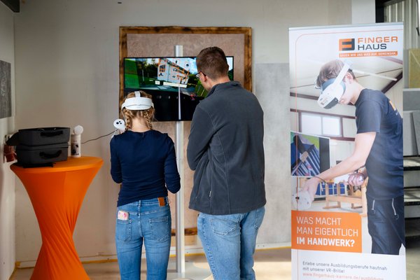 Zwei Personen mit VR-Brillen stehen vor einem Bildschirm und einem Banner mit dem Schriftzug 'Finger Haus' und der Frage 'Was macht man eigentlich im Handwerk'