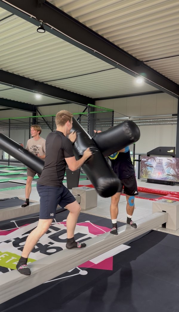 Zwei junge Männer stehen auf schmalen Balken und kämpfen spielerisch mit großen schwarzen Schaumstoffkeulen in einer Halle mit hoher Decke