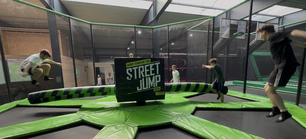 Drei Jugendliche springen auf einem grünen Trampolin mit schwarzem Mittelteil und der Aufschrift 'STREET JUMP' in einer Halle.