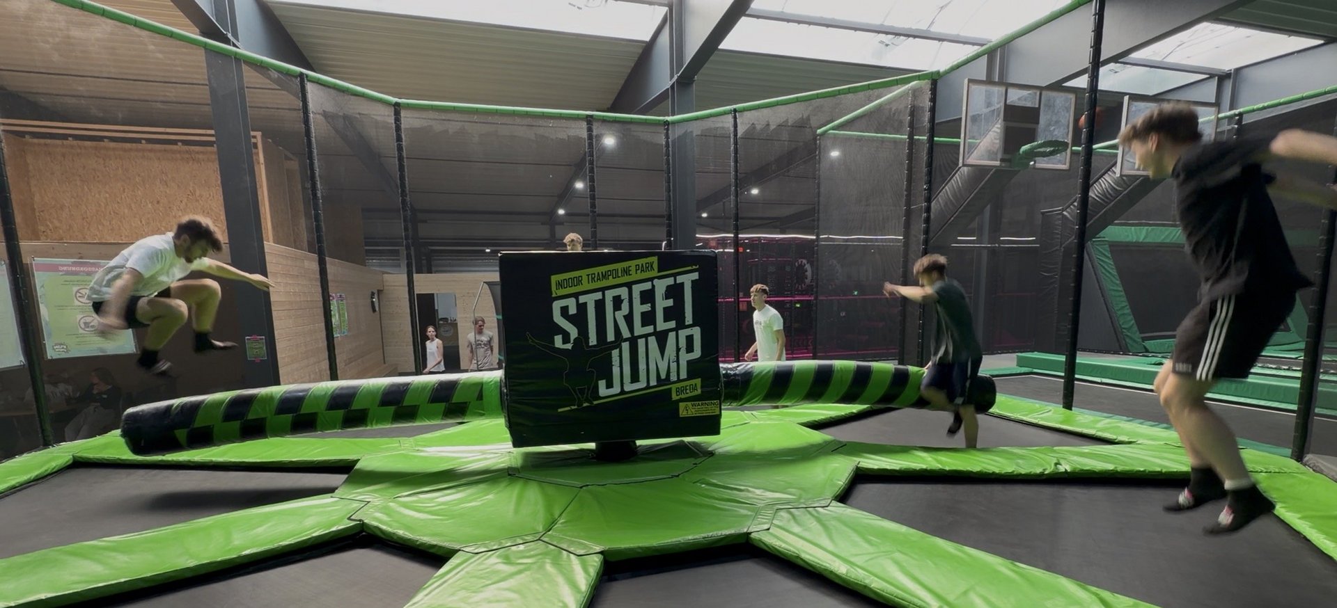 Drei Jugendliche springen auf einem grünen Trampolin mit schwarzem Mittelteil und der Aufschrift 'STREET JUMP' in einer Halle.