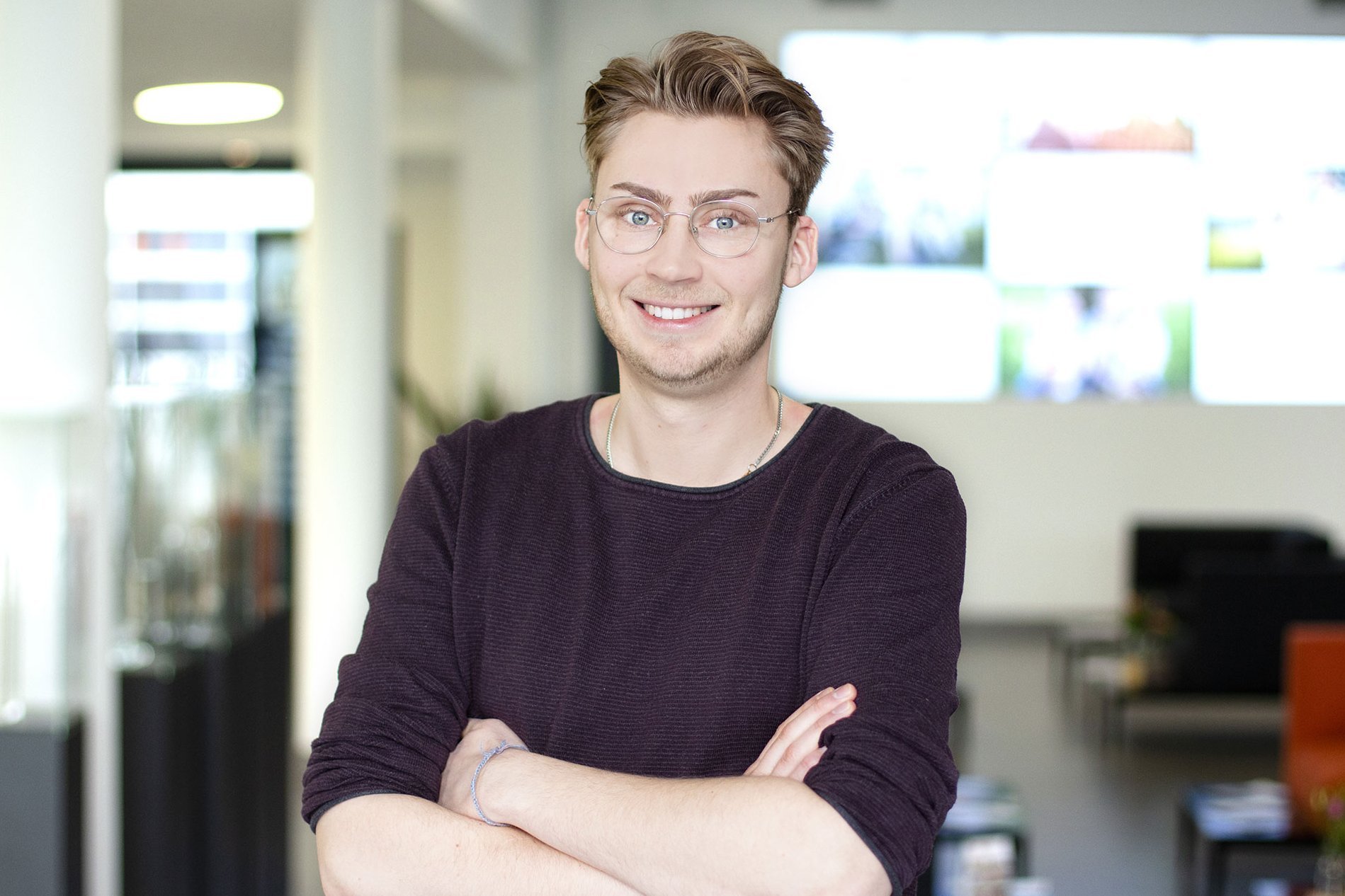 Junger Mann mit verschränkten Armen trägt dunkelroten Pullover und steht in einem modernen Büro mit Glaswänden und Schreibtischen im Hintergrund