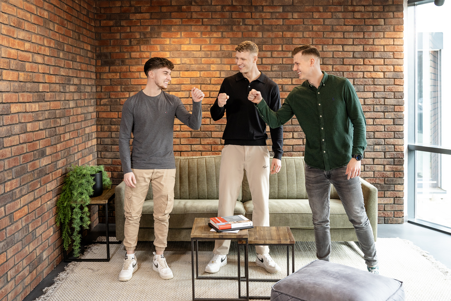 Drei junge Männer stehen in einem modernen Raum mit Ziegelsteinwand und führen eine Faustgruß-Geste aus, im Hintergrund ein Sofa und ein kleiner Tisch mit Büchern.